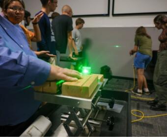 Person in a blue shirt uses a green laser on wooden blocks mounted on a metal track during a classroom demonstration.