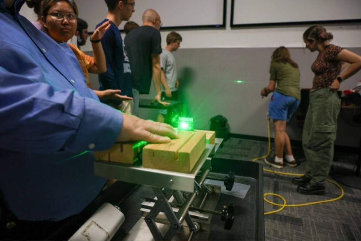 Person in a blue shirt uses a green laser on wooden blocks mounted on a metal track during a classroom demonstration.