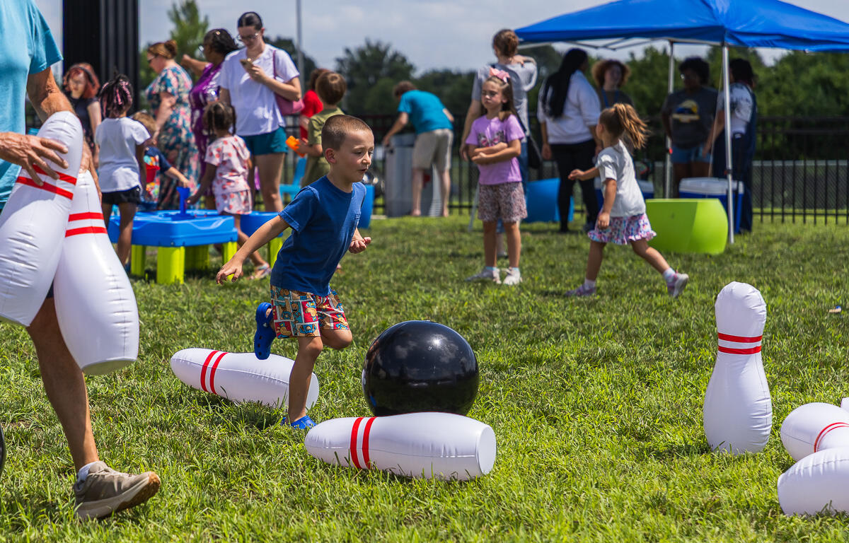 Young boy in a blue shirt runs toward a black bowling ball among large inflatable pins on a sunny grassy field at a kids' event.