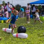 Young boy in a blue shirt runs toward a black bowling ball among large inflatable pins on a sunny grassy field at a kids' event.