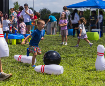 Young boy in a blue shirt runs toward a black bowling ball among large inflatable pins on a sunny grassy field at a kids' event.
