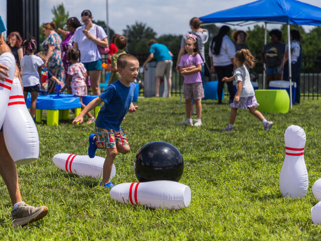 Young boy in a blue shirt runs toward a black bowling ball among large inflatable pins on a sunny grassy field at a kids' event.