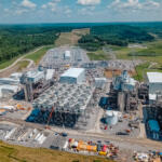 Aerial view of a large power plant complex with numerous circular cooling towers, cranes, and industrial buildings set in a rural, treed landscape.
