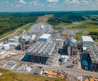 Aerial view of a large power plant complex with numerous circular cooling towers, cranes, and industrial buildings set in a rural, treed landscape.