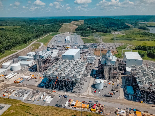 Aerial view of a large power plant complex with numerous circular cooling towers, cranes, and industrial buildings set in a rural, treed landscape.