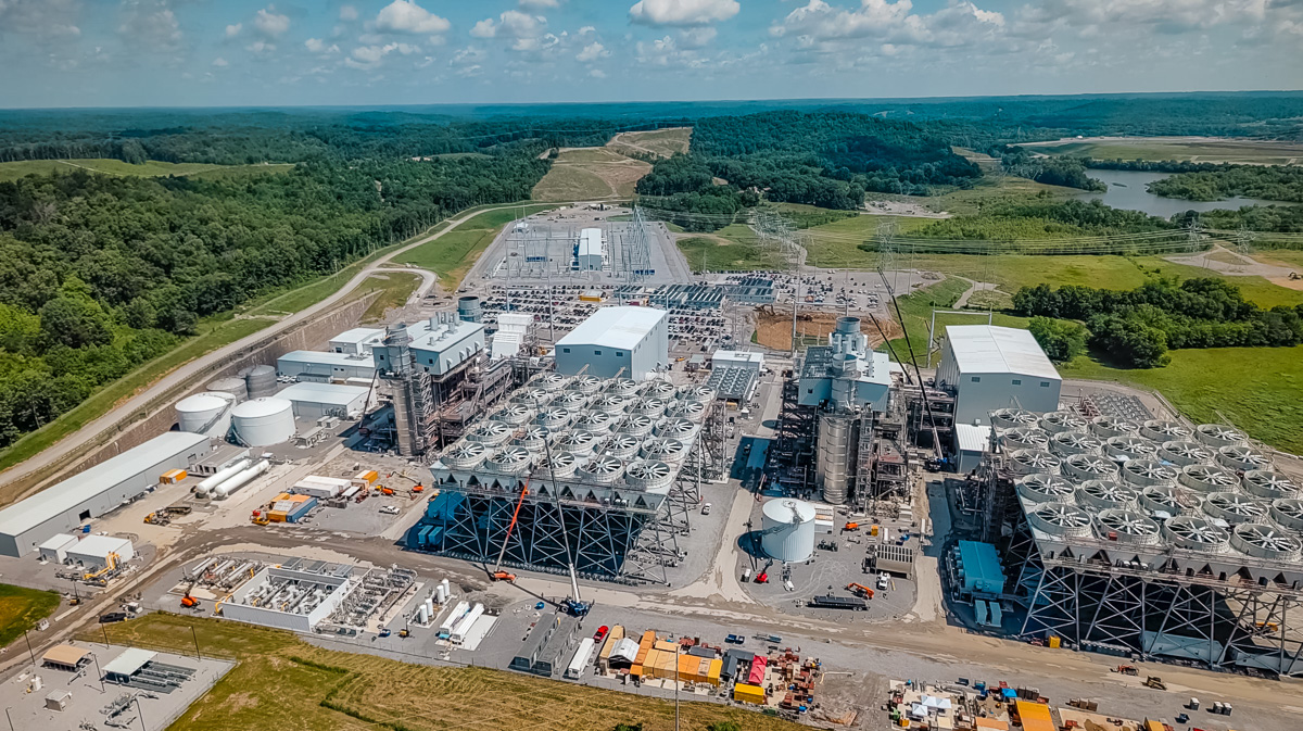Aerial view of a large power plant complex with numerous circular cooling towers, cranes, and industrial buildings set in a rural, treed landscape.