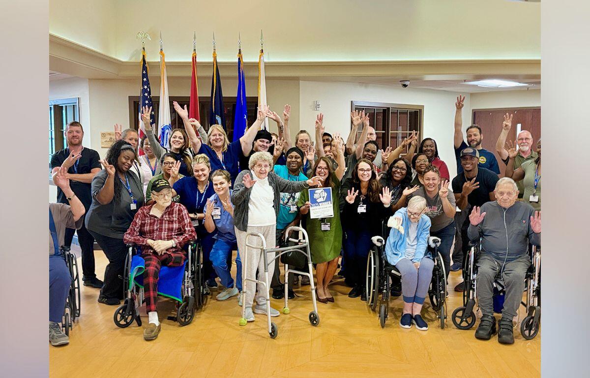 Group photo of a diverse community team and residents, many in wheelchairs, raising hands in a cheerful indoor gathering with flags in the background.