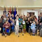Group photo of a diverse community team and residents, many in wheelchairs, raising hands in a cheerful indoor gathering with flags in the background.