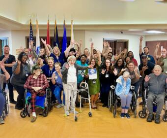 Group photo of a diverse community team and residents, many in wheelchairs, raising hands in a cheerful indoor gathering with flags in the background.