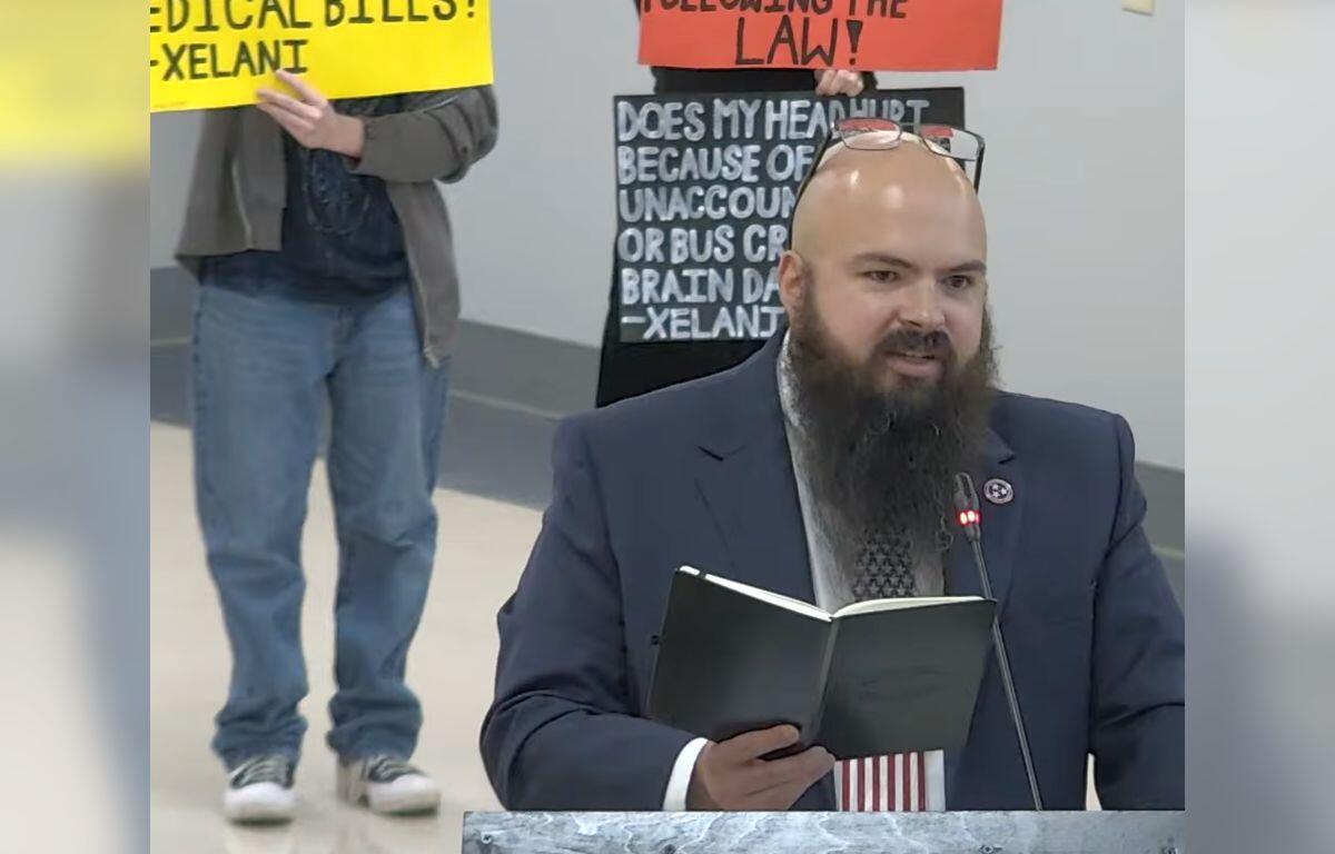 Bearded man in a suit at a podium reading a notebook, with protesters and bright signs in the background.
