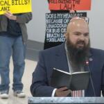 Bearded man in a suit at a podium reading a notebook, with protesters and bright signs in the background.