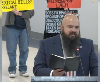 Bearded man in a suit at a podium reading a notebook, with protesters and bright signs in the background.