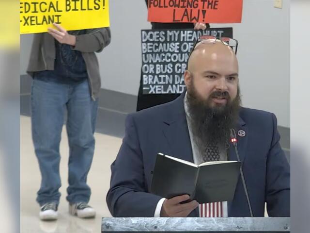 Bearded man in a suit at a podium reading a notebook, with protesters and bright signs in the background.