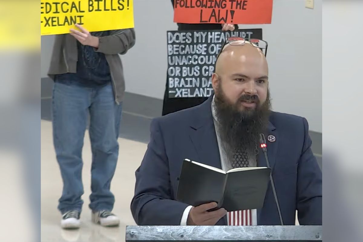 Bearded man in a suit at a podium reading a notebook, with protesters and bright signs in the background.