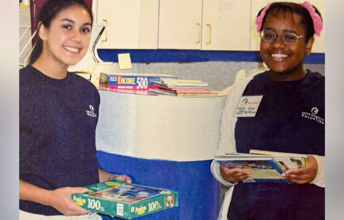 Two smiling young volunteers pose in a room with books, each holding puzzle or activity boxes.