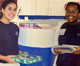 Two smiling young volunteers pose in a room with books, each holding puzzle or activity boxes.