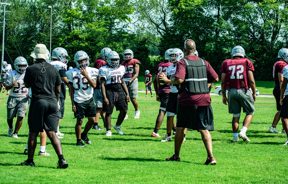 Football practice on a sunny field with players in white and maroon jerseys wearing helmets, listening to a coach in front.