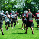 Football practice on a sunny field with players in white and maroon jerseys wearing helmets, listening to a coach in front.