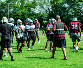 Football practice on a sunny field with players in white and maroon jerseys wearing helmets, listening to a coach in front.