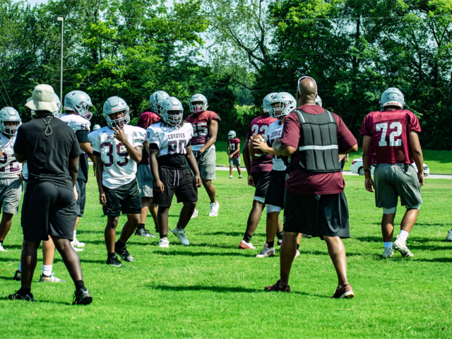 Football practice on a sunny field with players in white and maroon jerseys wearing helmets, listening to a coach in front.