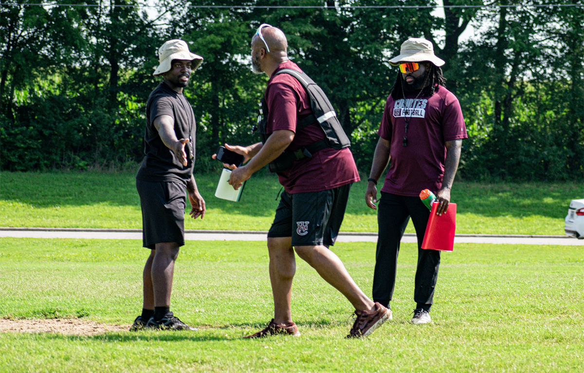 Three men on a grassy field during a sports practice; one extends a hand while another carries a water bottle and gear.