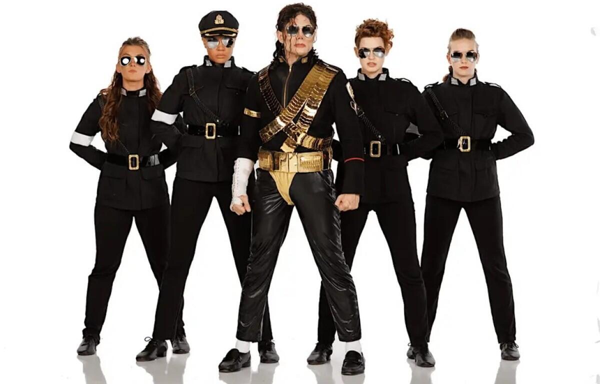 Five people stand in a line wearing black military-style outfits with belts and sunglasses; center performer wears gold accessories and sash.