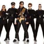Five people stand in a line wearing black military-style outfits with belts and sunglasses; center performer wears gold accessories and sash.