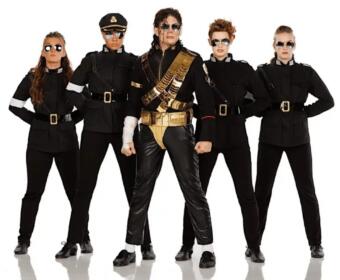 Five people stand in a line wearing black military-style outfits with belts and sunglasses; center performer wears gold accessories and sash.