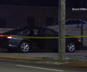 Nighttime street scene with a black sedan taped off by yellow caution tape; police cars visible in the background under dark sky.