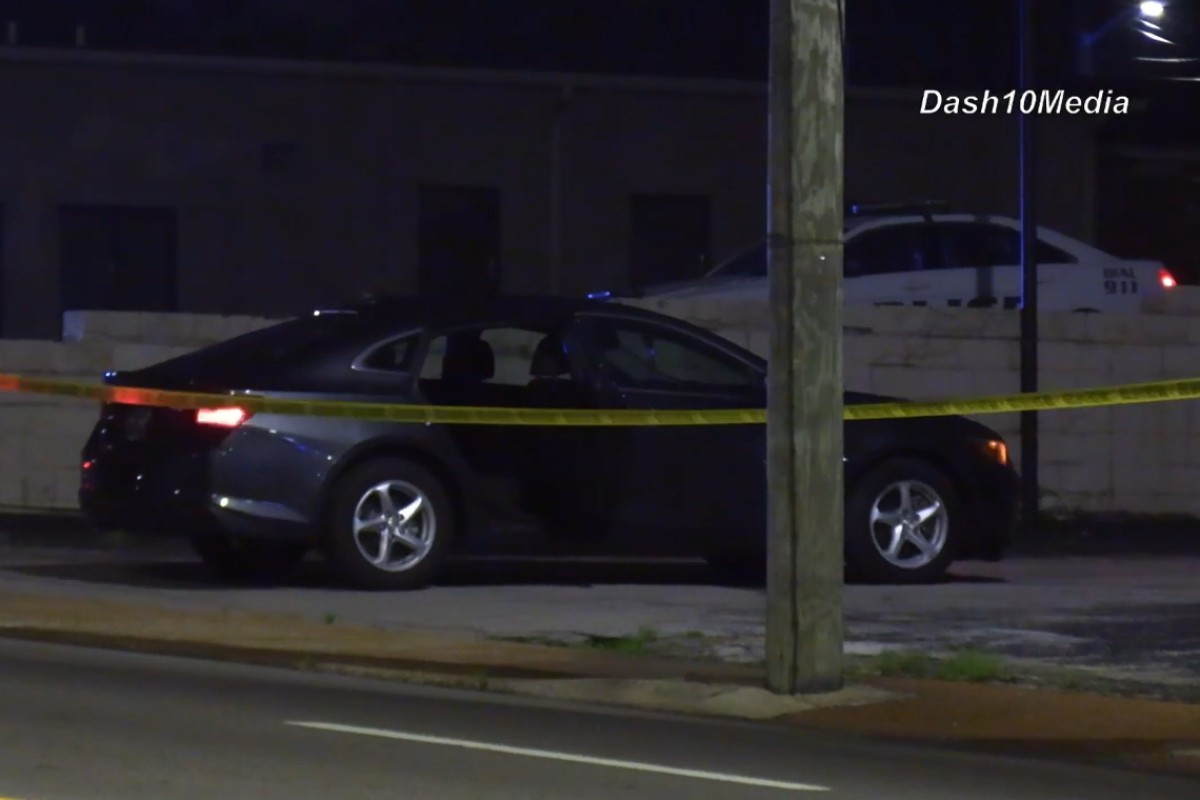 Nighttime street scene with a black sedan taped off by yellow caution tape; police cars visible in the background under dark sky.
